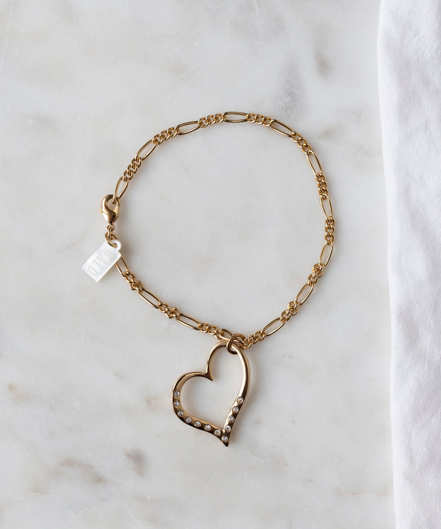The WALD Berlin Valentines Day special sketch heart bracelet features a heart-shaped pendant with sparkling Swarovski stones, shown on a white marble surface beside a white cloth.