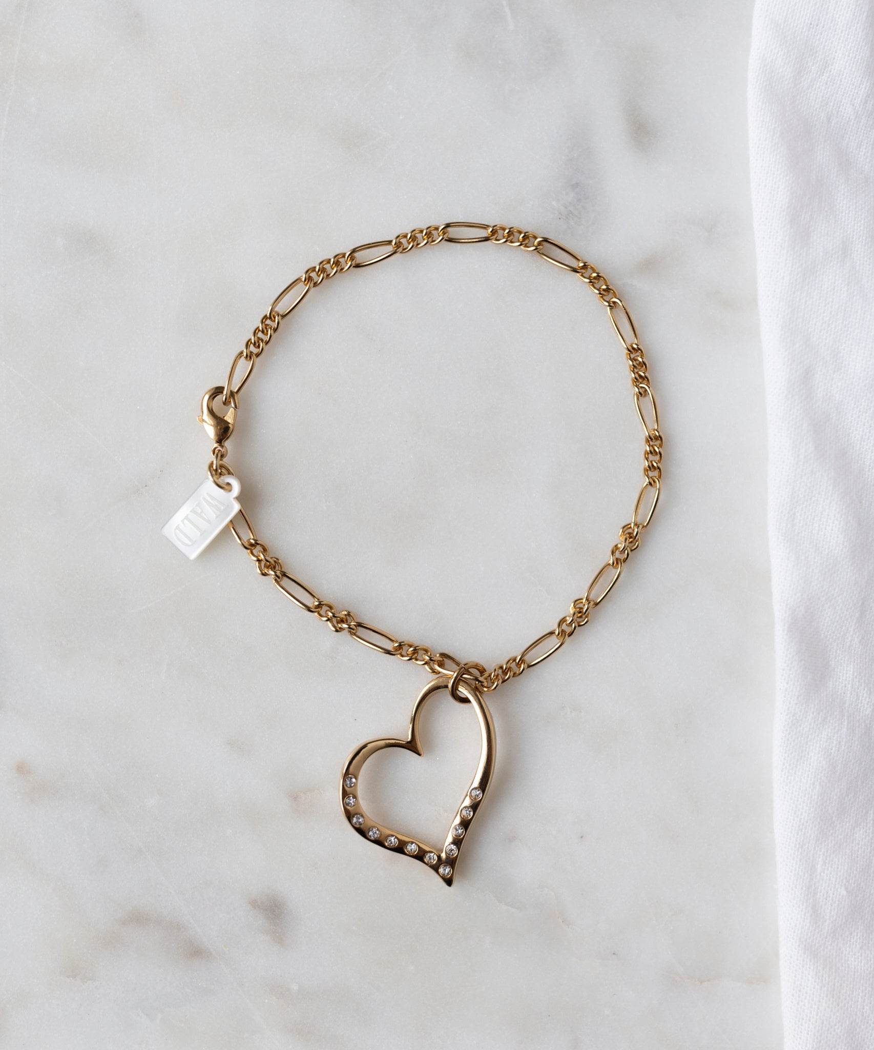 The WALD Berlin Valentines Day special sketch heart bracelet features a heart-shaped pendant with sparkling Swarovski stones, shown on a white marble surface beside a white cloth.