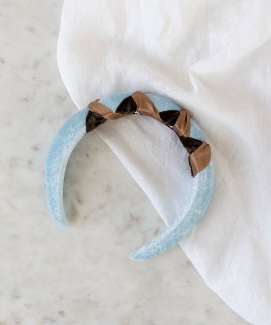 The SAMPLE SALE Headband Knot Light Blue by WALD Berlin, handmade in Germany, features twisted brown and black satin ribbon accents. It is displayed on a white marble surface next to a white cloth.