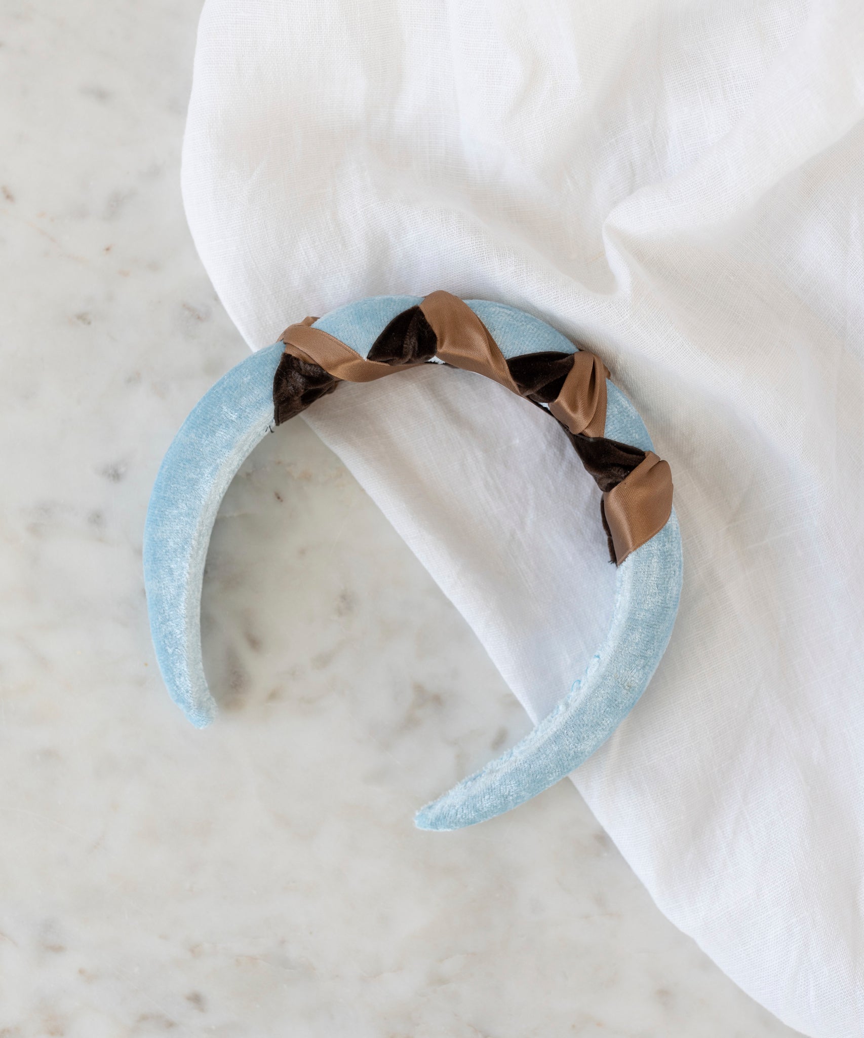 The SAMPLE SALE Headband Knot Light Blue by WALD Berlin, handmade in Germany, features twisted brown and black satin ribbon accents. It is displayed on a white marble surface next to a white cloth.