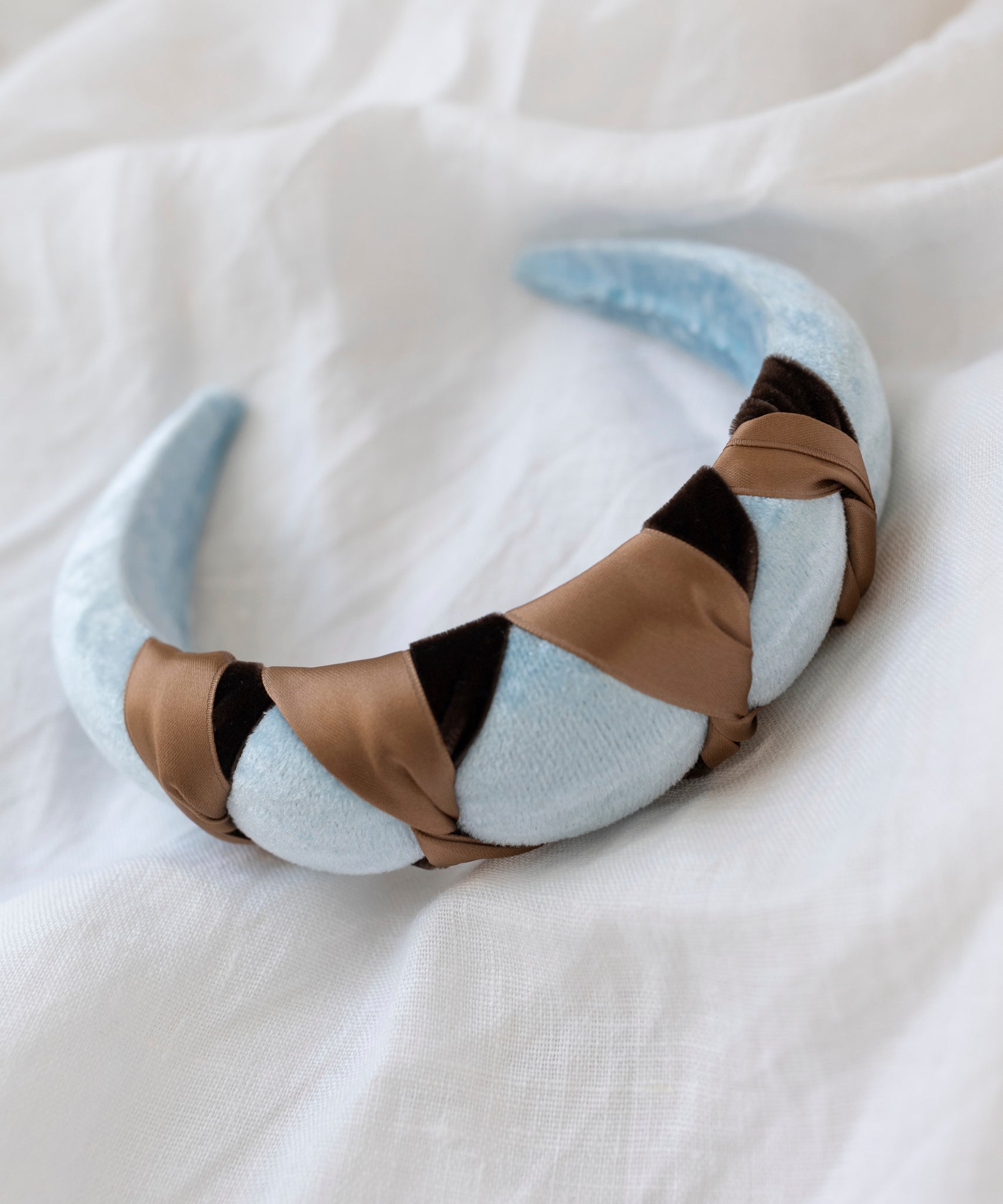 The SAMPLE SALE Headband Knot Light Blue by WALD Berlin is handmade in Germany, featuring a light blue velvety base with a brown and black satin braided design, displayed on a white fabric surface.