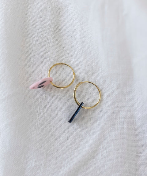 The WALD Berlin SAMPLE SALE From Another Mother Gold-Blue Hoops, featuring two charming gold hoop earrings adorned with a pink disc and a black bar, are elegantly displayed on a soft white fabric background. These earrings are crafted from high-quality bio polyester and are made in Germany.