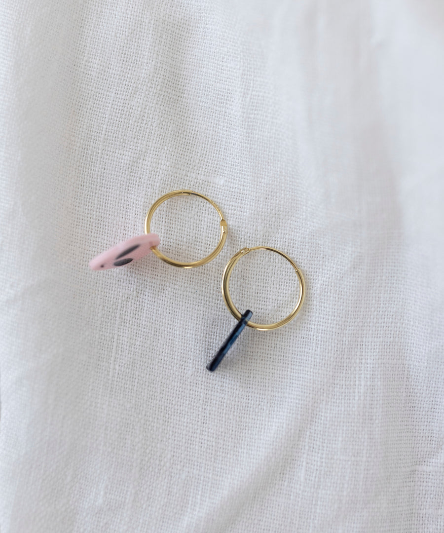 The WALD Berlin SAMPLE SALE From Another Mother Gold-Blue Hoops, featuring two charming gold hoop earrings adorned with a pink disc and a black bar, are elegantly displayed on a soft white fabric background. These earrings are crafted from high-quality bio polyester and are made in Germany.