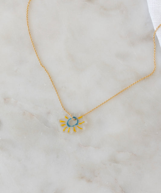 The SAMPLE SALE Daisy Necklace WALD World Transparent by WALD Berlin features a sun-shaped pendant with a blue center and yellow rays on a gold chain, crafted from recycled gold and silver, displayed on light marble.