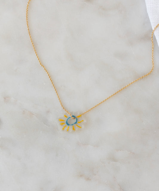 The SAMPLE SALE Daisy Necklace WALD World Transparent by WALD Berlin features a sun-shaped pendant with a blue center and yellow rays on a gold chain, crafted from recycled gold and silver, displayed on light marble.