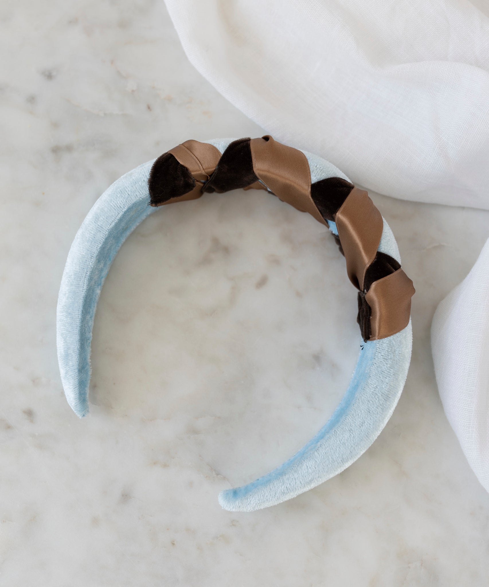 The SAMPLE SALE Headband Knot Light Blue by WALD Berlin, handmade in Germany, features a light blue velvet design with brown and black braided satin detail, displayed on white marble beside white fabric.