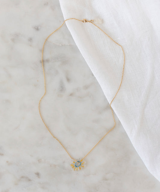 The SAMPLE SALE Daisy Necklace WALD World Transparent by WALD Berlin, crafted from recycled gold and silver with a daisy-shaped pendant, rests on light marble beside white textured fabric.