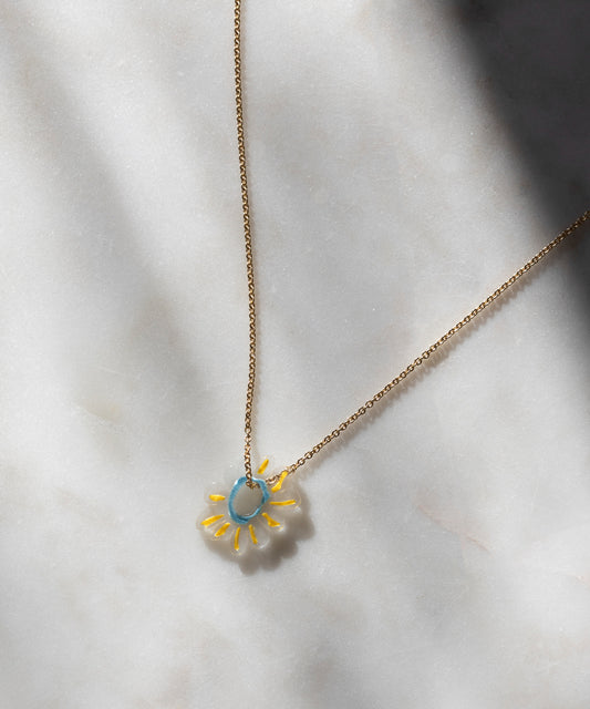 The SAMPLE SALE Daisy Necklace by WALD Berlin features a daisy-shaped pendant with a blue center and yellow and white petals, crafted from recycled gold and silver, elegantly displayed on a light marble surface.