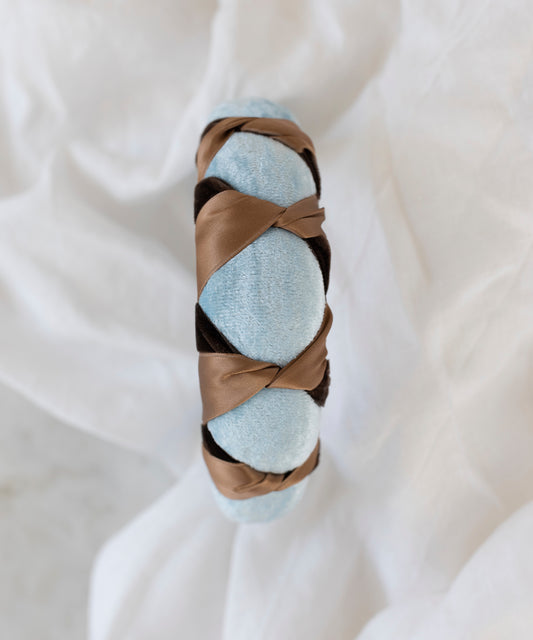 The SAMPLE SALE Headband Knot Light Blue by WALD Berlin, handmade in Germany, showcases light blue velvet with a brown satin ribbon in a crisscross design, displayed on a white cloth background.