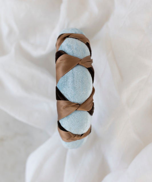 The SAMPLE SALE Headband Knot Light Blue by WALD Berlin, handmade in Germany, showcases light blue velvet with a brown satin ribbon in a crisscross design, displayed on a white cloth background.