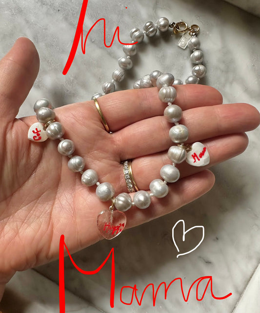 A hand holds the WALD Berlin Limited HI MAMA Pearl Necklace, featuring real sweet water pearls and heart-shaped charms. "Hi Mamma" appears in red over the image as the necklace rests on a marble surface.