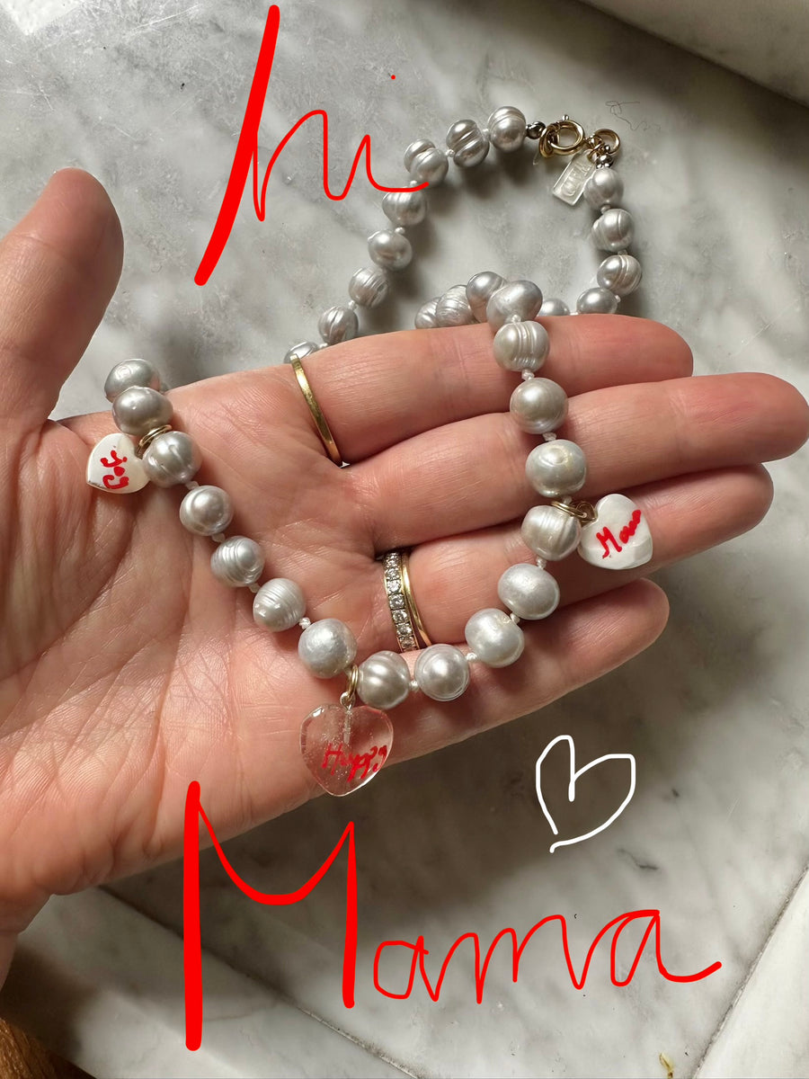 A hand holds the WALD Berlin Limited HI MAMA Pearl Necklace, featuring real sweet water pearls and heart-shaped charms. "Hi Mamma" appears in red over the image as the necklace rests on a marble surface.