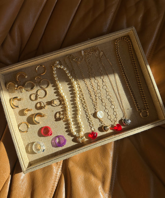 A jewelry tray on brown leather holds gold earrings, rings, and necklaces with pearls, beads, and heart pendants—partially lit by sunlight beside a chic surprise WALD Berlin Bag from WALD Berlin.