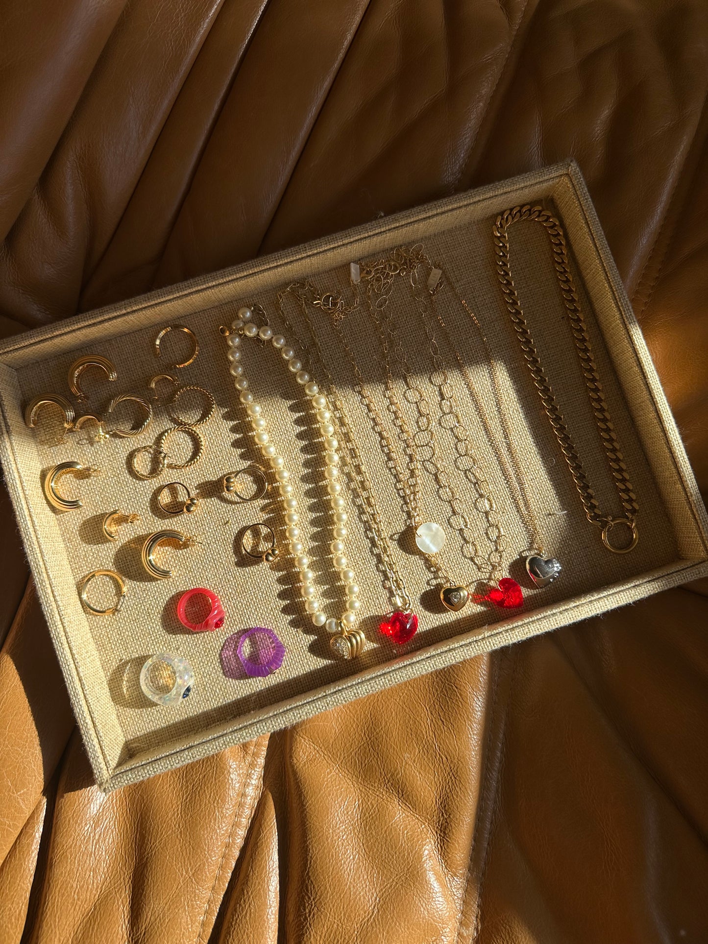 A jewelry tray on brown leather holds gold earrings, rings, and necklaces with pearls, beads, and heart pendants—partially lit by sunlight beside a chic surprise WALD Berlin Bag from WALD Berlin.