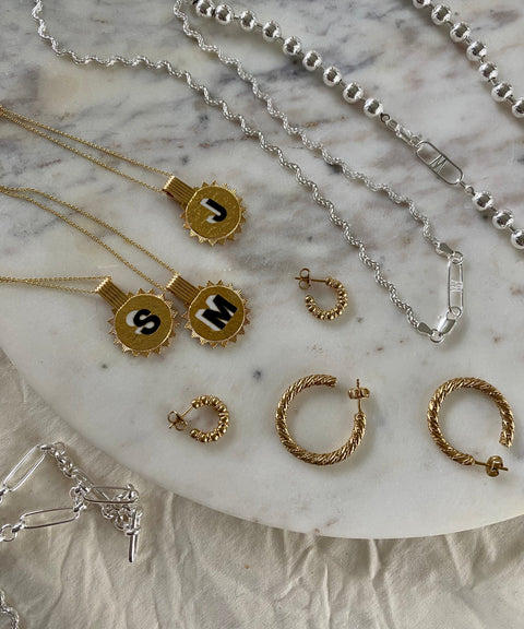 The Naomi Bracelet Silver by WALD Berlin is displayed on a marble surface alongside initial necklaces, textured hoop earrings, and various recycled gold and silver chain styles.