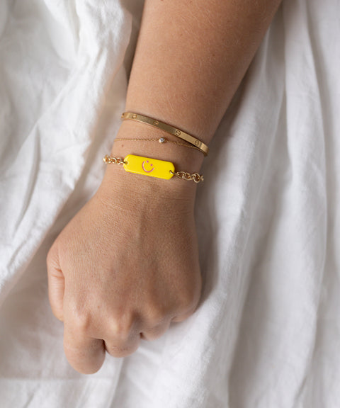 A hand wears three bracelets: an 18k gold plated bangle, a delicate chain with bead, and the WALD Berlin SAMPLE SALE Happy ID Yellow bracelet exclusive with a smiley face tag. White fabric background. Made in Germany.
