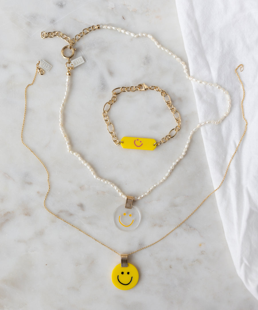 On a marble surface: WALD Berlin SAMPLE SALE Happy ID Yellow bracelet exclusive with pearls and 18k gold plating, plus two necklaces—one pearl and gold with a clear smiley pendant, one gold with a yellow smiley pendant.