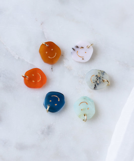 Five Smilie Dude Gemstone Pendants by WALD Berlin, each featuring colorful real gemstones with engraved smiley faces and 18k gold loops, are displayed on white marble. Crafted in Germany, pendants are sold without a chain.