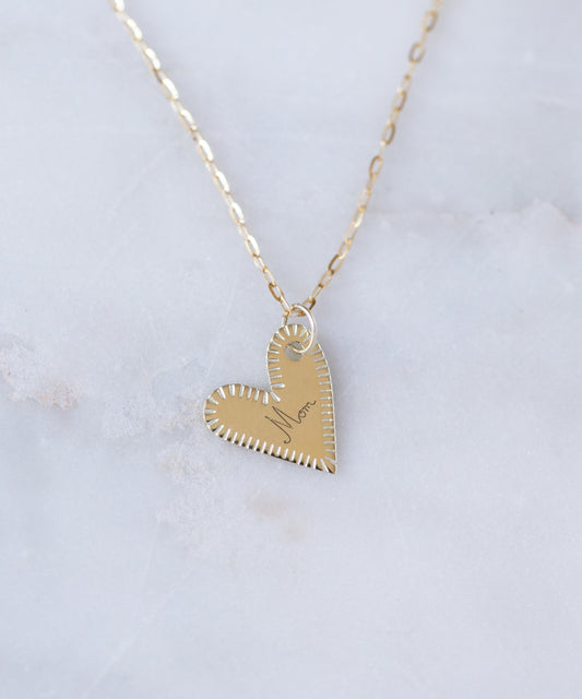 The WALD Berlin Be My Lover Heart 18k Pendant Without Chain, engraved with "Mom," is elegantly displayed on a light marble surface. Thoughtfully crafted in Germany, this stunning jewelry piece celebrates heartfelt connections.