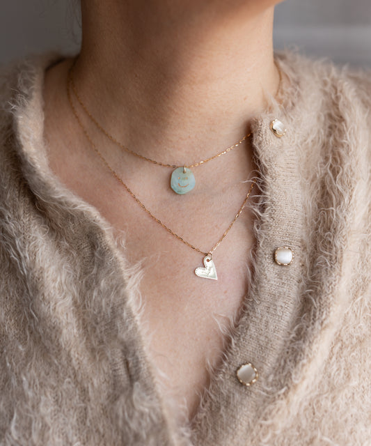 A person wearing a beige textured sweater with pearl buttons and two layered gold necklaces, featuring the WALD Berlin Be My Lover Heart 18k Pendant Without Chain and a round pendant, highlights the elegance of engraved jewelry.
