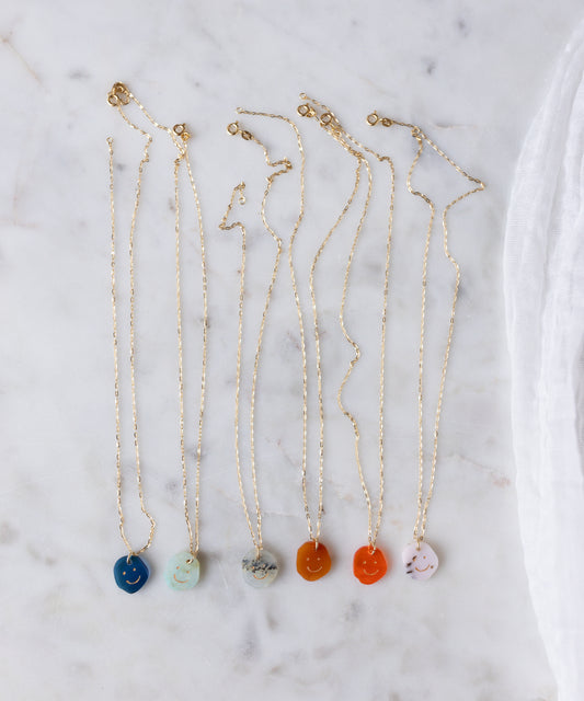 Six WALD Berlin Smilie Dude Gemstone Pendants Without Chain—blue, turquoise, white, gray, orange, and red—are displayed on white marble. Each 18k gold necklace features a playful yet elegant colorful smiley face design.