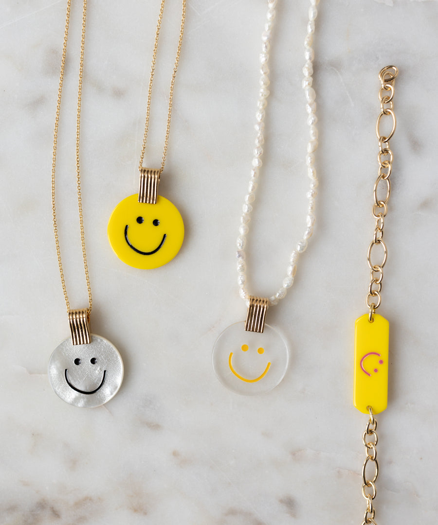 Four jewelry pieces on marble: two 18k gold plated chains with smiley pendants, a pearl necklace with clear smiley, and the WALD Berlin SAMPLE SALE Happy ID Yellow bracelet exclusive.