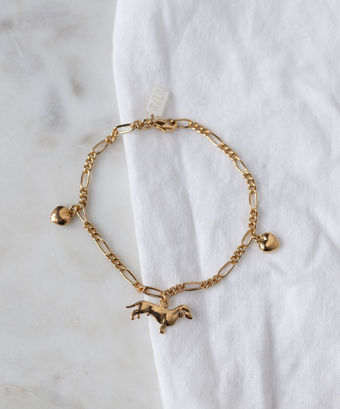 The WALD Berlin SAMPLE SALE Charm Bracelet Z Edition, 18k gold plated with three charms (heart, horse, heart), is displayed on a white fabric surface.