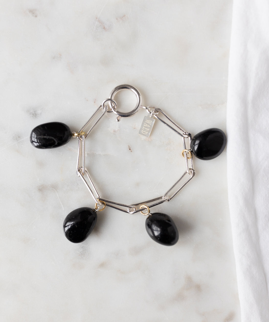 The WALD Berlin Collection Noir Ashley Drop Silver Bracelet features four black oval stone charms and a small rectangular tag, crafted from recycled silver and displayed on white marble.