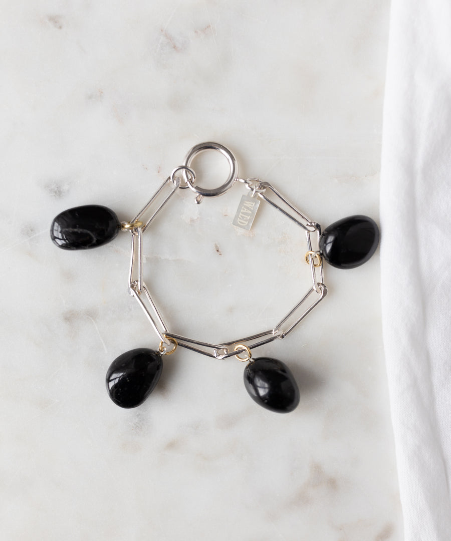 The WALD Berlin Collection Noir Ashley Drop Silver Bracelet features four black oval stone charms and a small rectangular tag, crafted from recycled silver and displayed on white marble.