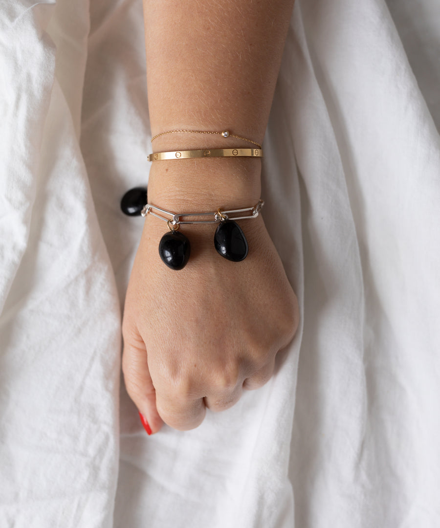 A hand wearing a gold bangle, a chain bracelet, and WALD Berlin’s Collection Noir Ashley Drop Silver Bracelet with three black stone charms rests on white fabric.