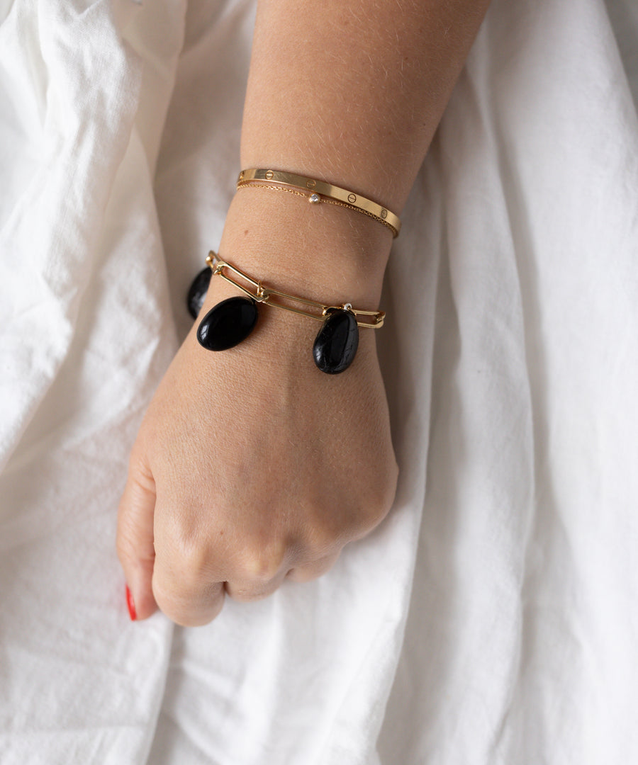 A hand with a gold bangle and the WALD Berlin Collection Noir Ashley Drop Gold Bracelet featuring three black stone charms rests on white fabric.