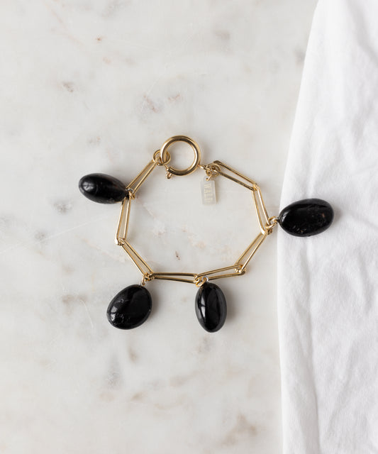 The WALD Berlin Collection Noir Ashley Drop Gold Bracelet features four large black oval stone charms, crafted in recycled gold and 18k gold plating, displayed on a white marble surface beside a white cloth.