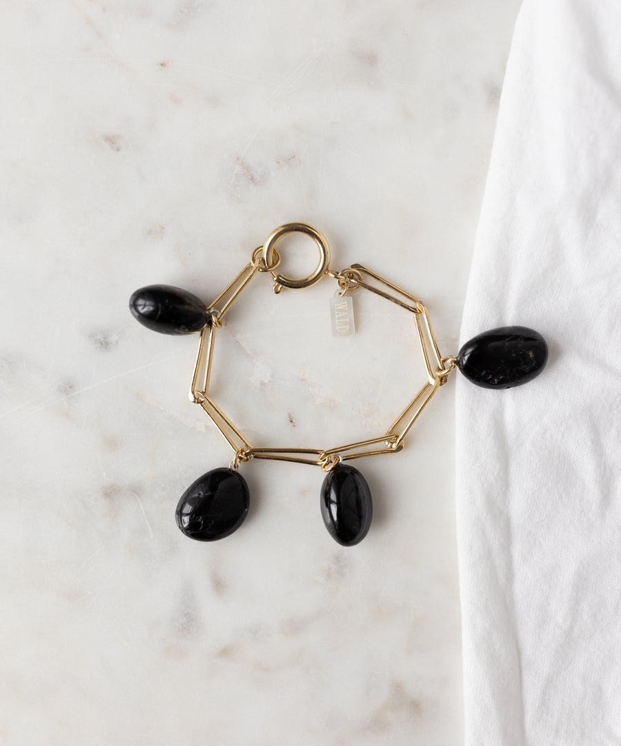 The WALD Berlin Collection Noir Ashley Drop Gold Bracelet features four large black oval stone charms, crafted in recycled gold and 18k gold plating, displayed on a white marble surface beside a white cloth.