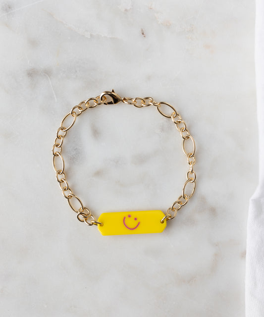The WALD Berlin Happy ID Yellow bracelet exclusive features an 18k gold plated chain and a yellow rectangular charm with a pink smiley face, shown on a white marble surface.