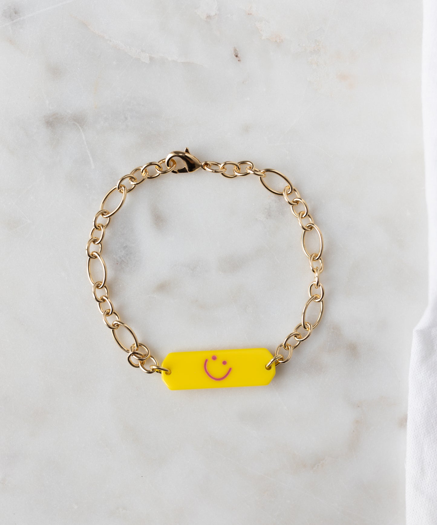 The WALD Berlin Happy ID Yellow bracelet exclusive features an 18k gold plated chain and a yellow rectangular charm with a pink smiley face, shown on a white marble surface.