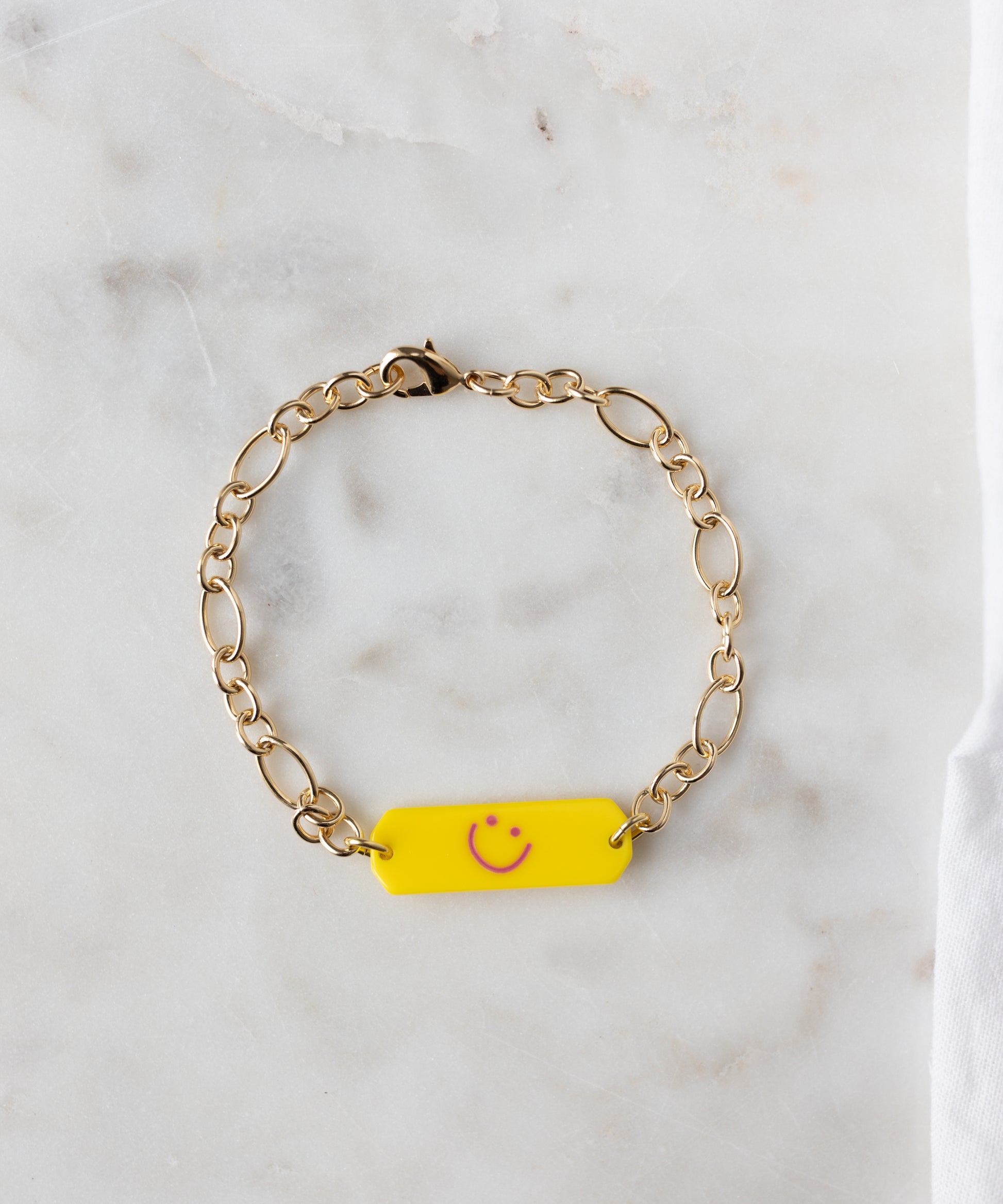 The WALD Berlin Happy ID Yellow bracelet exclusive features an 18k gold plated chain and a yellow rectangular charm with a pink smiley face, shown on a white marble surface.