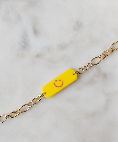 The WALD Berlin SAMPLE SALE Happy ID Yellow bracelet exclusive features an 18k gold-plated chain with a rectangular yellow tag and pink smiley face, displayed on a marble surface.