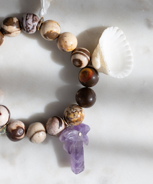 The WALD Berlin Mexicana La Bamba Bracelet, handmade in Germany, showcases brown and beige stones, a white seashell, and a purple carved stone charm, displayed on a white marble surface.