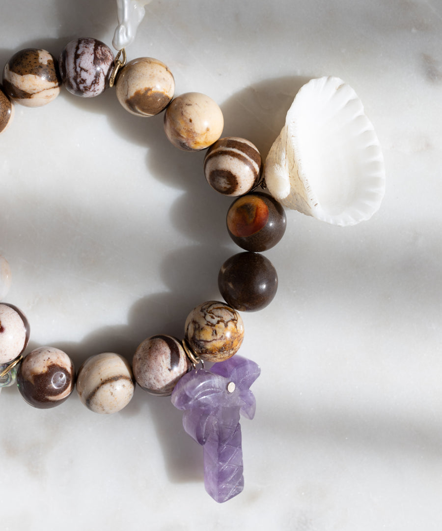 The WALD Berlin Mexicana La Bamba Bracelet, handmade in Germany, showcases brown and beige stones, a white seashell, and a purple carved stone charm, displayed on a white marble surface.