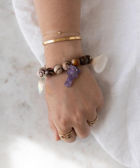 A hand adorned with WALD Berlin's Mexicana La Bamba Bracelet—featuring a purple stone, shell, and leaf charm—rests on white fabric alongside gold rings and thin gold bracelets. Handmade in Germany.