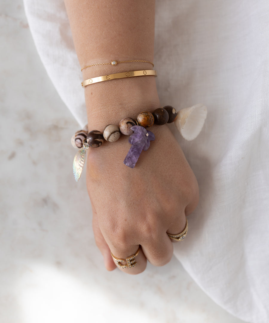A hand adorned with WALD Berlin's Mexicana La Bamba Bracelet—featuring a purple stone, shell, and leaf charm—rests on white fabric alongside gold rings and thin gold bracelets. Handmade in Germany.