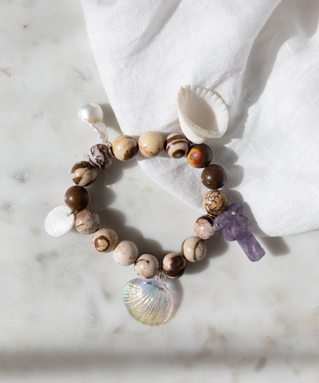 WALD Berlin's Mexicana La Bamba Bracelet, featuring picture jasper beads, a purple crystal, and three shell charms, is displayed on marble beside a white cloth and a small white seashell.