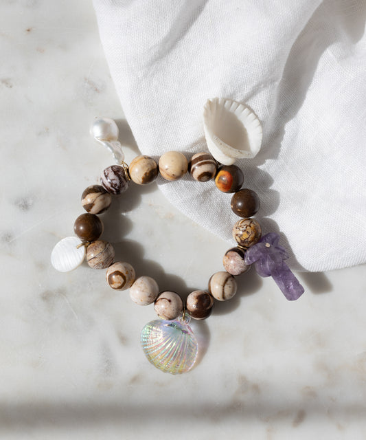 WALD Berlin's Mexicana La Bamba Bracelet, featuring picture jasper beads, a purple crystal, and three shell charms, is displayed on marble beside a white cloth and a small white seashell.