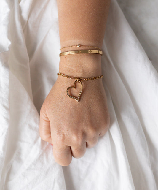 A hand rests on white fabric, wearing three 18k gold-plated bracelets from WALD Berlin, including the Valentines Day special sketch heart bracelet and another embellished with sparkling Swarovski stones.
