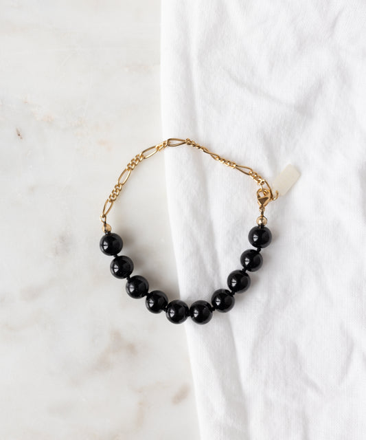 The WALD Berlin Collection Noir Pas De Deux Black Bracelet, adorned with seven black beads, is displayed on a white marble surface beside a white cloth.