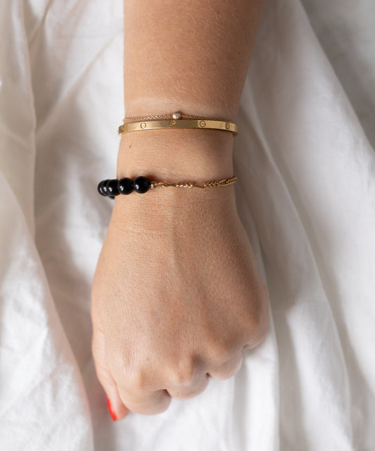 A hand rests on white fabric, adorned with an 18k gold plated bracelet, a thin gold chain, and the Collection Noir Pas De Deux Black Bracelet by WALD Berlin.