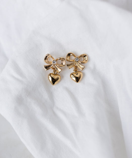 WALD Berlin's Valentines Day special bow heart earrings—gold-plated brass with bow designs and dangling heart charms—are shown on a white fabric background. Perfect as heart earrings for Valentine's Day.
