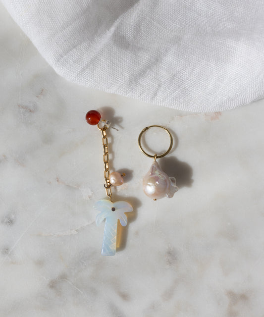 The Mexicana Mariachi Earrings Pearl by WALD Berlin are mismatched gold earrings with pearls, red bead, chain, and palm tree charm, displayed on marble. Handmade in Germany. A piece of white fabric is visible in the corner.