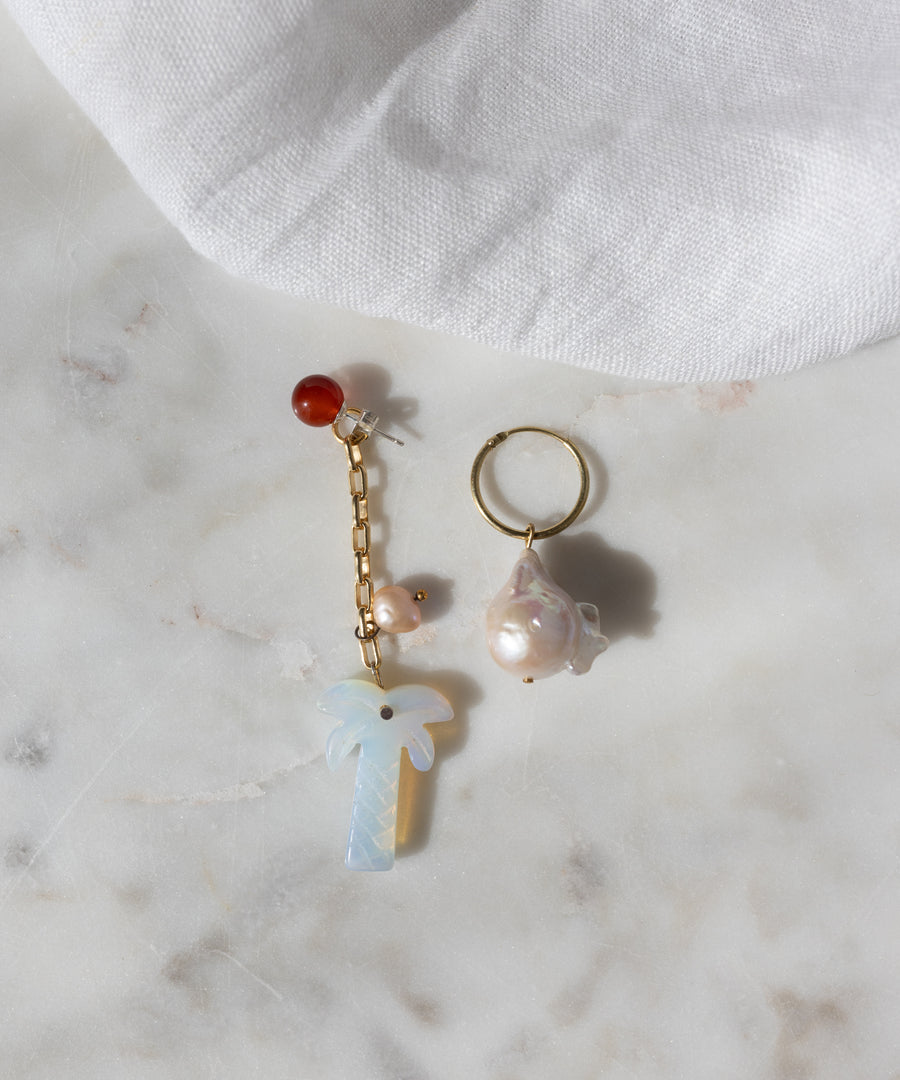 The Mexicana Mariachi Earrings Pearl by WALD Berlin are mismatched gold earrings with pearls, red bead, chain, and palm tree charm, displayed on marble. Handmade in Germany. A piece of white fabric is visible in the corner.