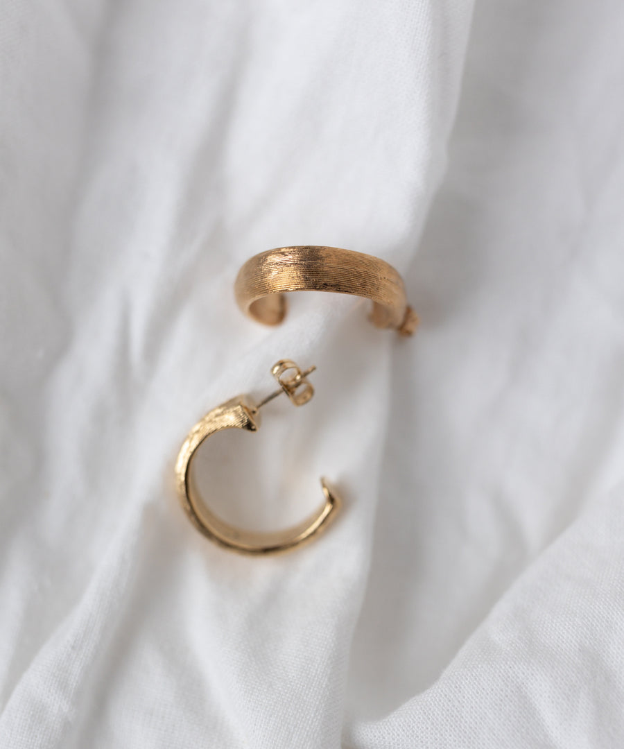 The WALD Berlin SAMPLE SALE Pop Classy Hoop Gold earrings, featuring an 18k gold plated textured finish, are displayed on a white fabric background.